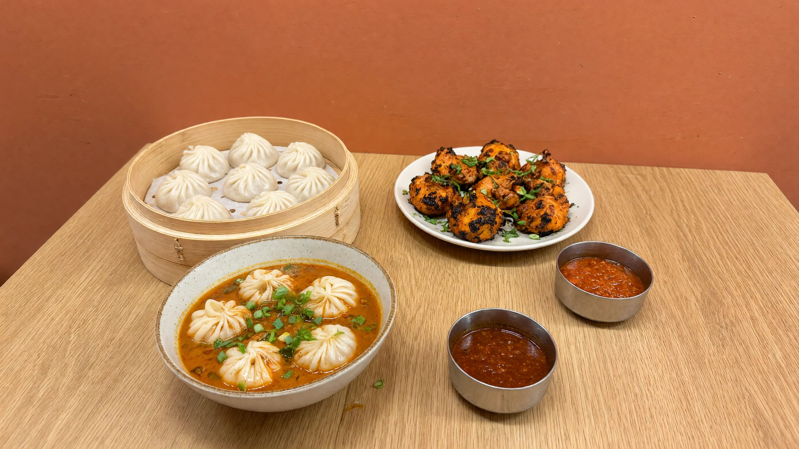 Steamed momos in a bamboo steamer, tandoori momos, jhol momo and small bowls of achar on a wooden table